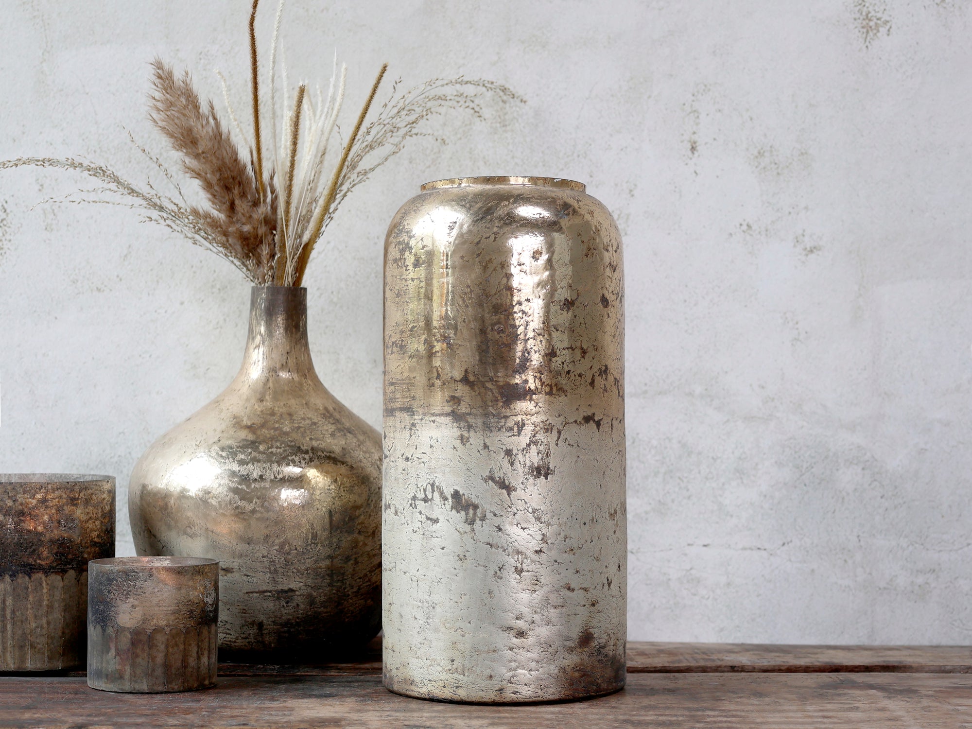 tall cylindrical decorative vase in a gold and bronze colour sat on a wooden surface against a neutral wall